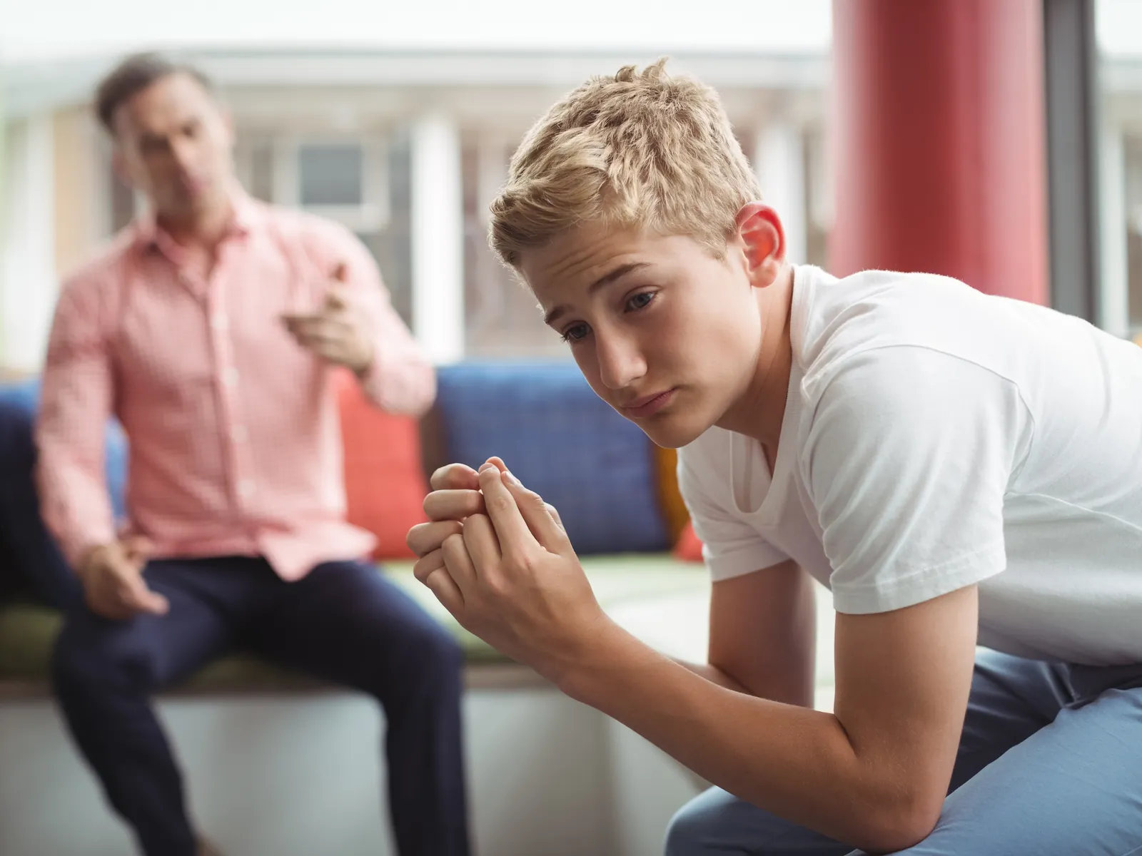 Adolescente con gesto preocupado mientras su padre le habla al fondo, distancia en la comunicación familiar