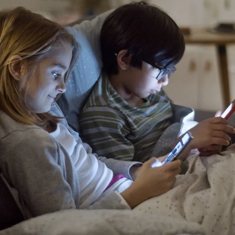 Niños con el móvil antes de dormir Niña y niño tapados con una manta en el sofá, usando el móvil por la noche